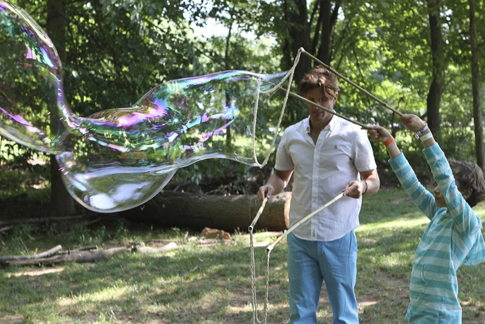 Make memories bigger with BubbleStix giant bubble kits A dad and his child pulling a long iridescent giant bubble across a shady backyard with BubbleStix
