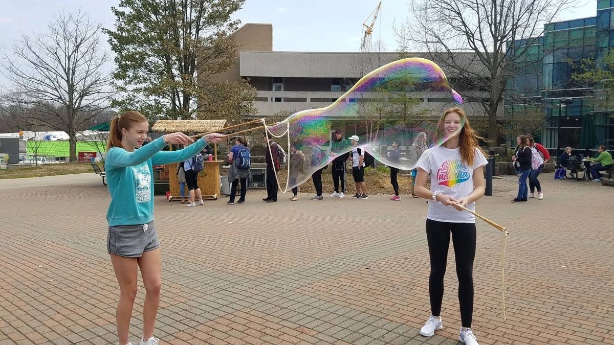 Two college students making a massive iridescent giant bubble together with BubbleStix on a campus plaza