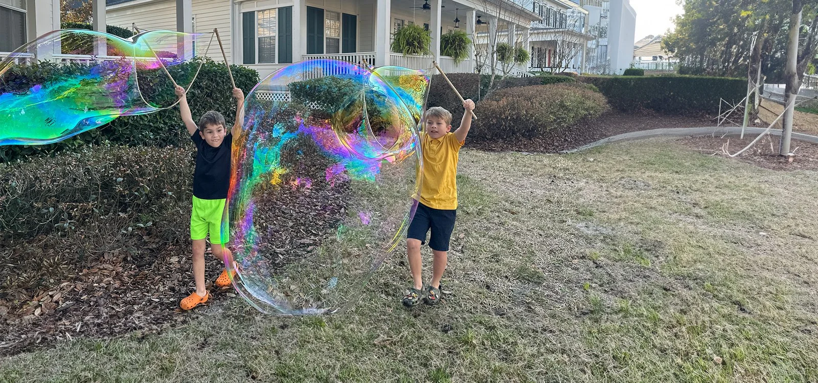 Two boys making a giant iridescent bubble together with the BubbleStix Double Pack in a front yard