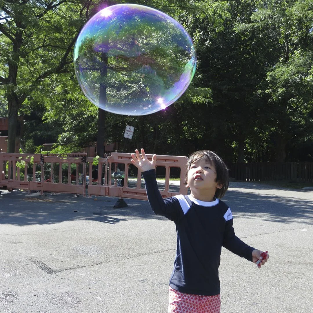 Chasing giant bubbles made with BubbleStix A young boy reaching up toward a giant iridescent bubble made with BubbleStix