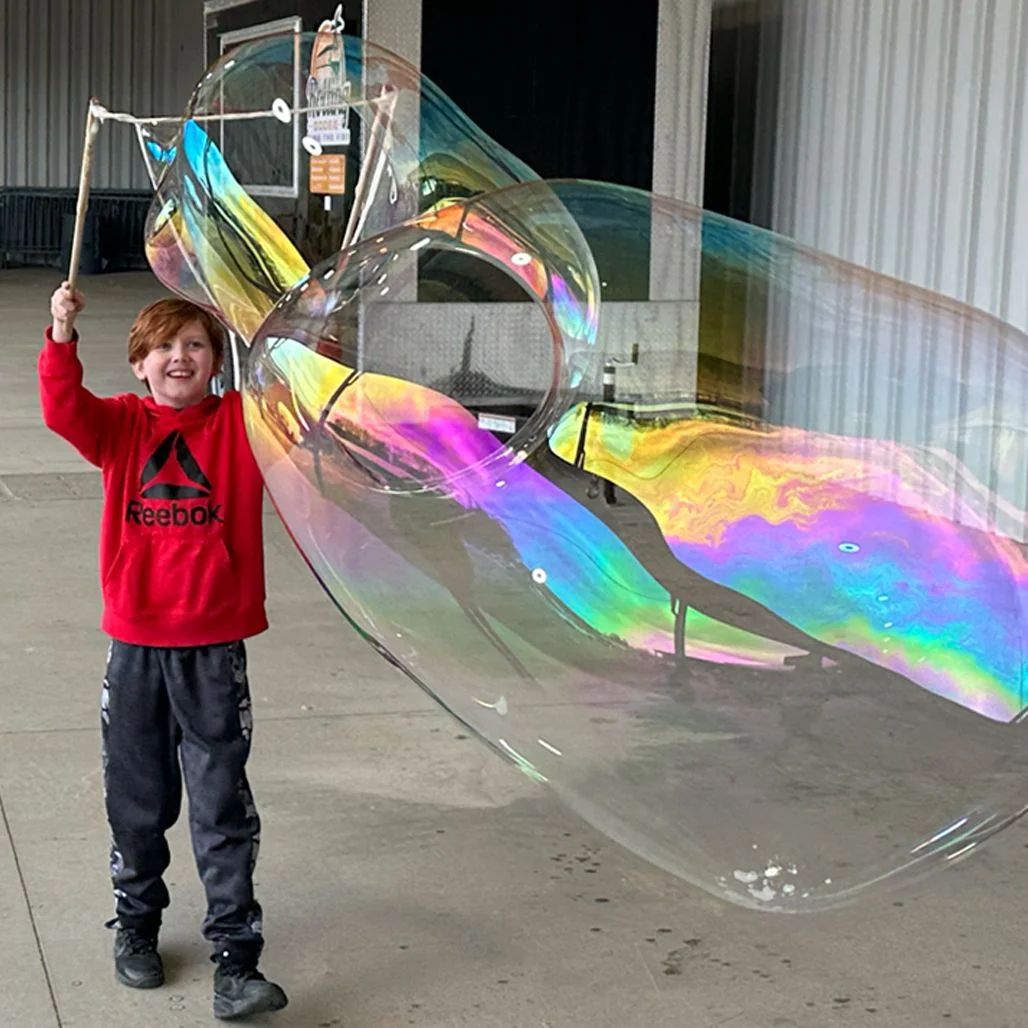 Screen-free outdoor fun with BubbleStix giant bubbles A boy in a red hoodie making a huge iridescent giant bubble outdoors with BubbleStix