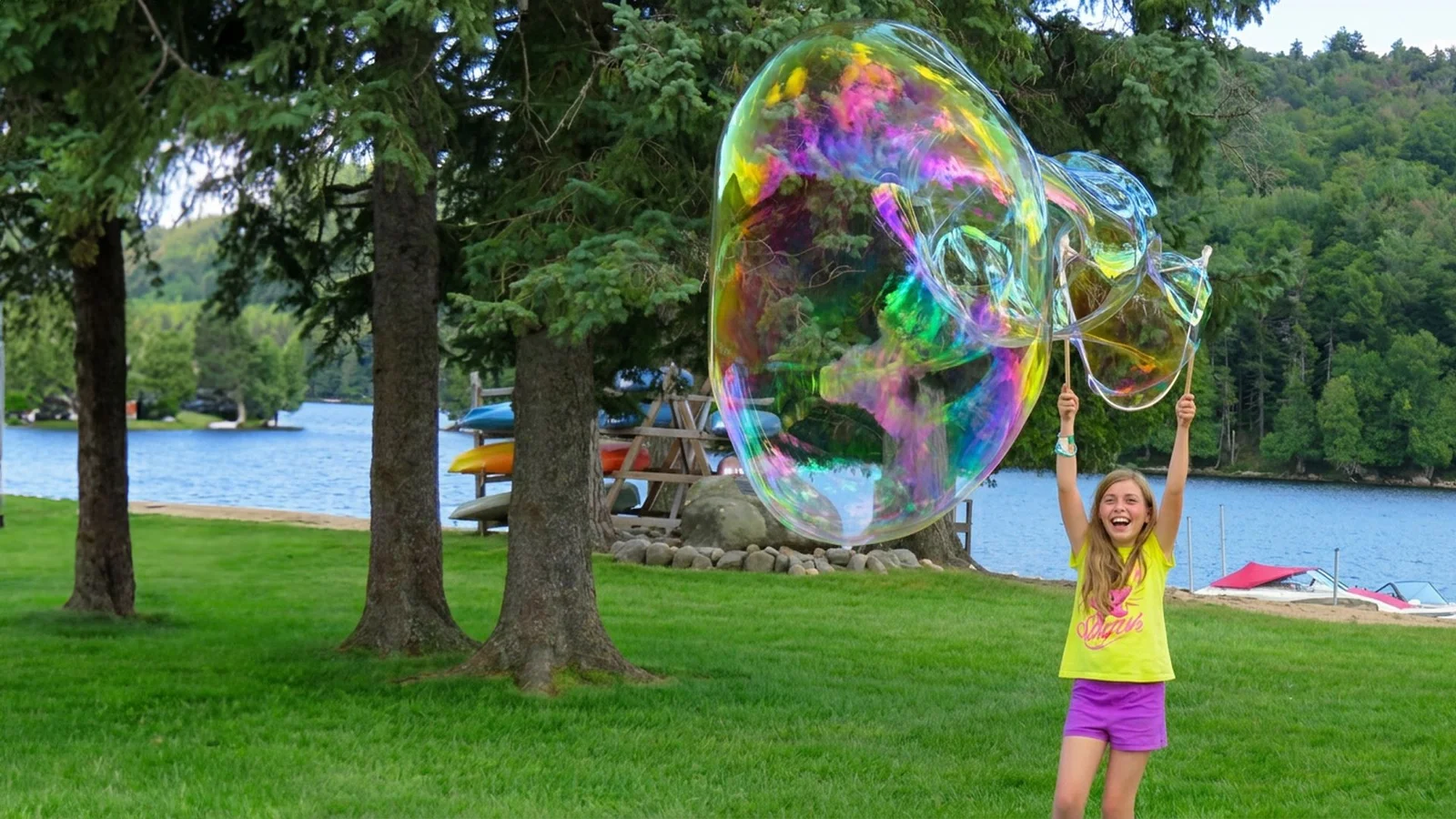 A girl making a huge iridescent giant bubble beside a lake with BubbleStix