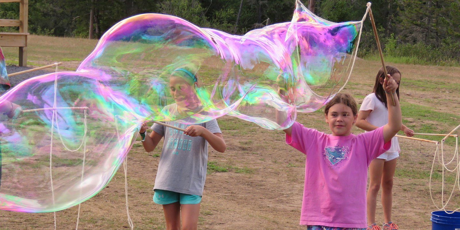 Two girls making an enormous iridescent giant bubble outdoors with handcrafted wooden BubbleStix