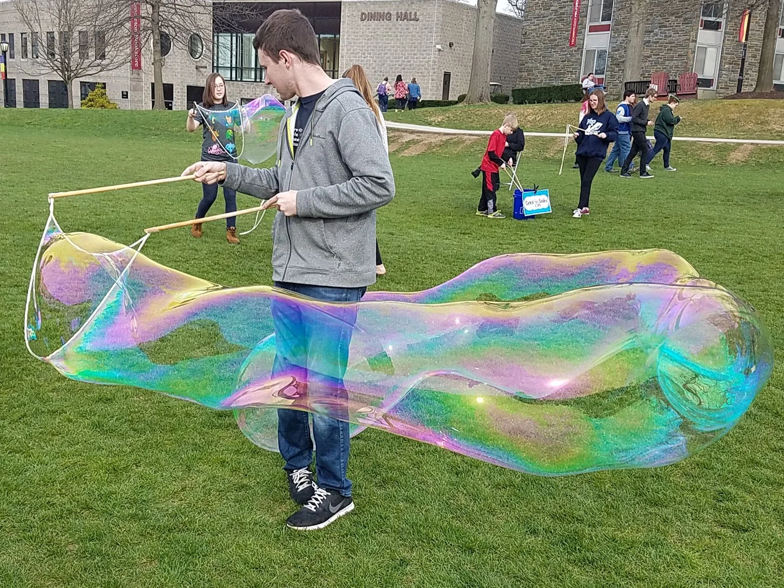BubbleStix review — Marcus T., Glenside, Pennsylvania A college student pulling a massive rainbow giant bubble across a campus lawn with BubbleStix