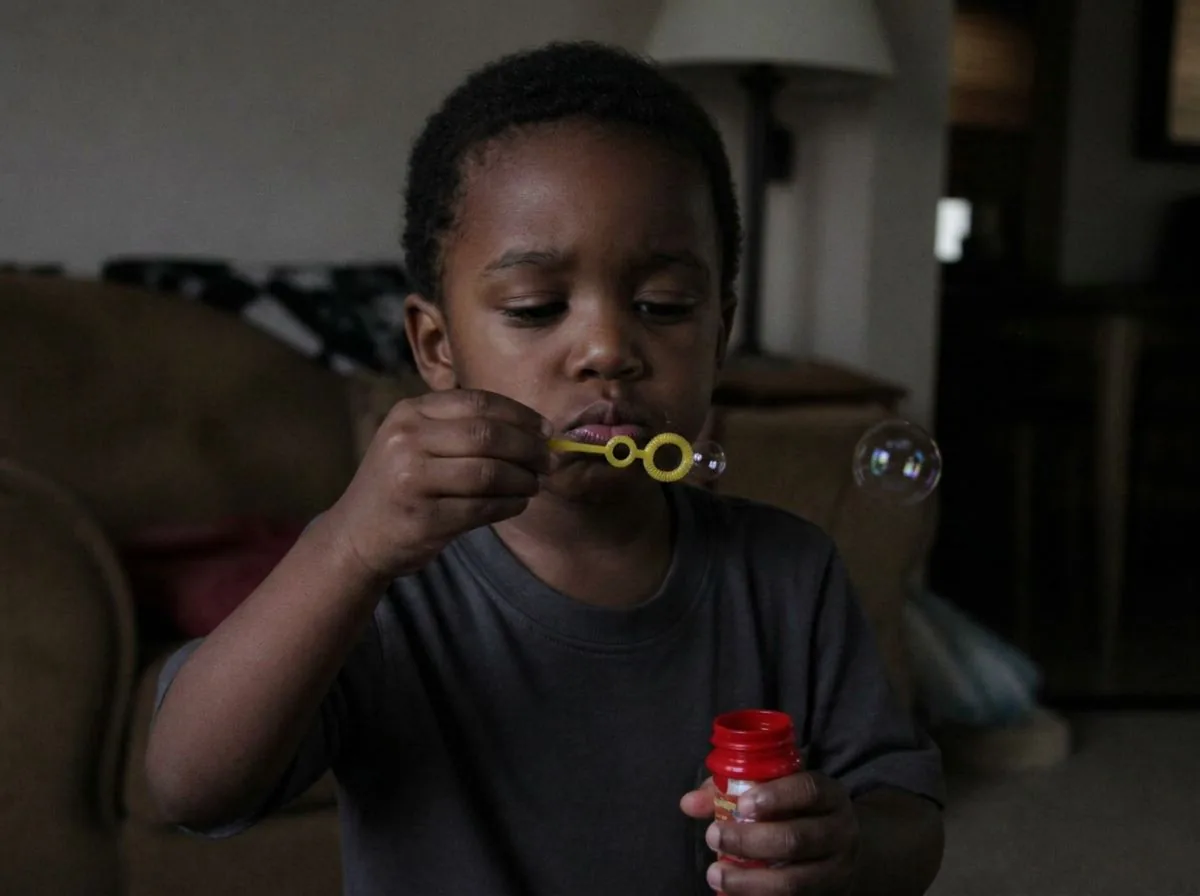 A child indoors blowing a small bubble from a plastic wand, with a bottle of store-bought bubble solution in his other hand