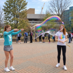 Two friends making a long giant bubble on a campus plaza with BubbleStix wooden bubble wands