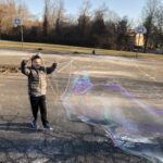 Boy making a long giant bubble with handcrafted BubbleStix wooden bubble sticks on an outdoor basketball court